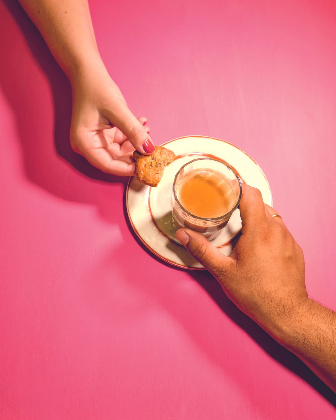 Zwei Hände teilen sich eine Tasse authentischen indischen Chai von Karma Food mit einem Keks auf einem pinken Tisch