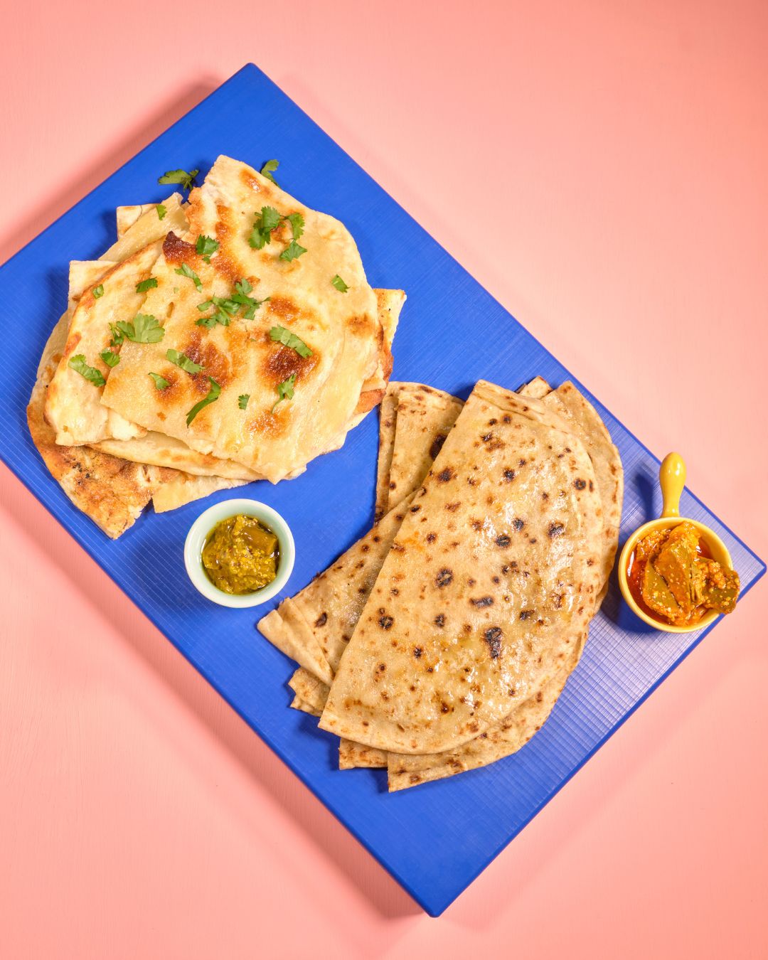 Zwei Sorten indisches Fladenbrot – fluffiges Naan mit Koriander und dünnes Roti – serviert auf einem blauen Brett mit zwei Schälchen Achar (indischem Pickle), auf rosa Hintergrund.