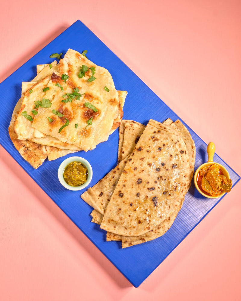 Zwei Sorten indisches Fladenbrot – fluffiges Naan mit Koriander und dünnes Roti – serviert auf einem blauen Brett mit zwei Schälchen Achar (indischem Pickle), auf rosa Hintergrund.