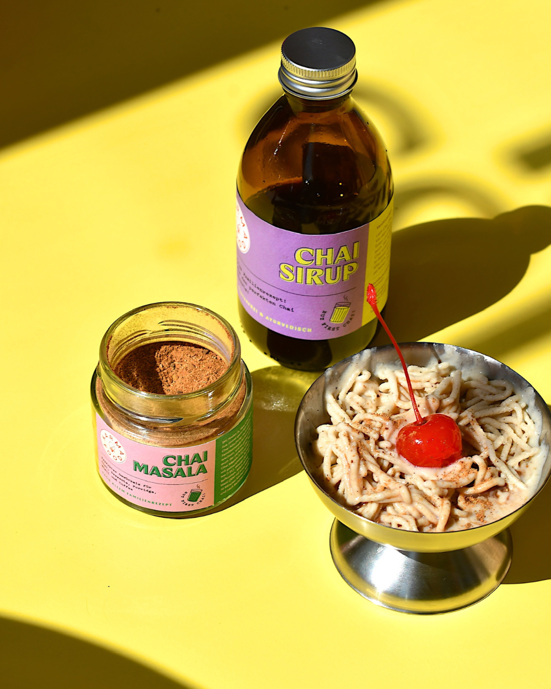 Vanille Spaghetti Eis mit roter Cocktailkirsche in Metallschale, daneben Glas mit Chai Masala Pulver und Flasche Chai Sirup auf gelbem Hintergrund