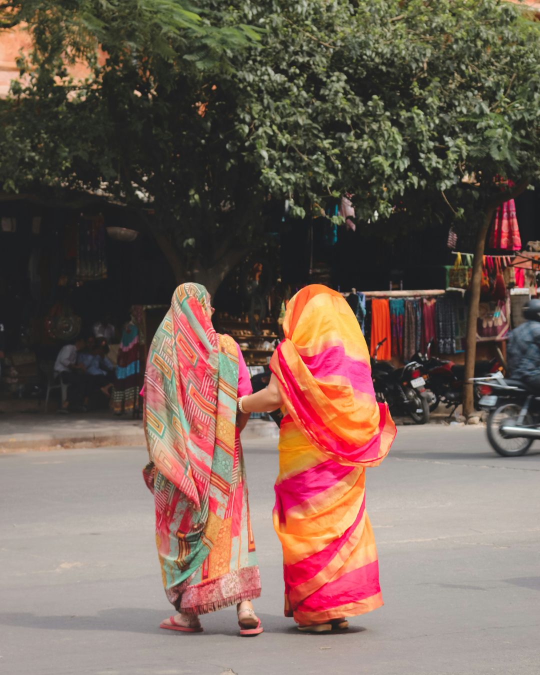 Zwei Frauen in bunten Saris spazieren Arm in Arm über eine Straße in Indien, im Hintergrund Marktstände und Motorräder