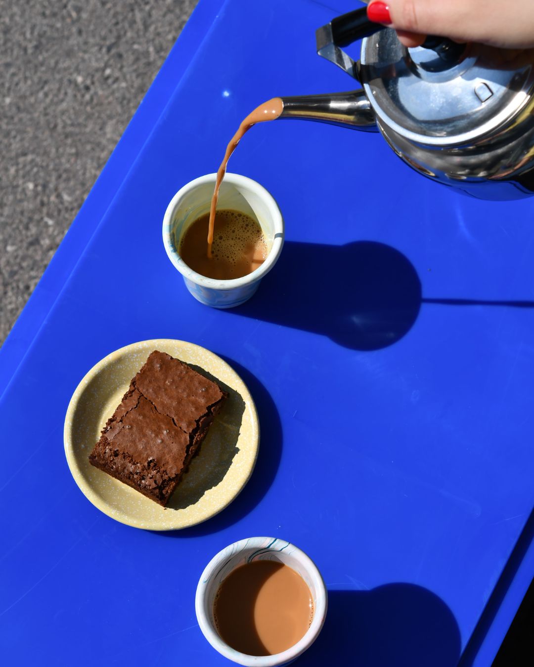 Hand gießt Chai aus einer Metallkanne in einen Becher auf einem blauen Tisch. Daneben ein Teller mit Brownie und ein zweiter Becher Chai. Szene in hellem Sonnenlicht.