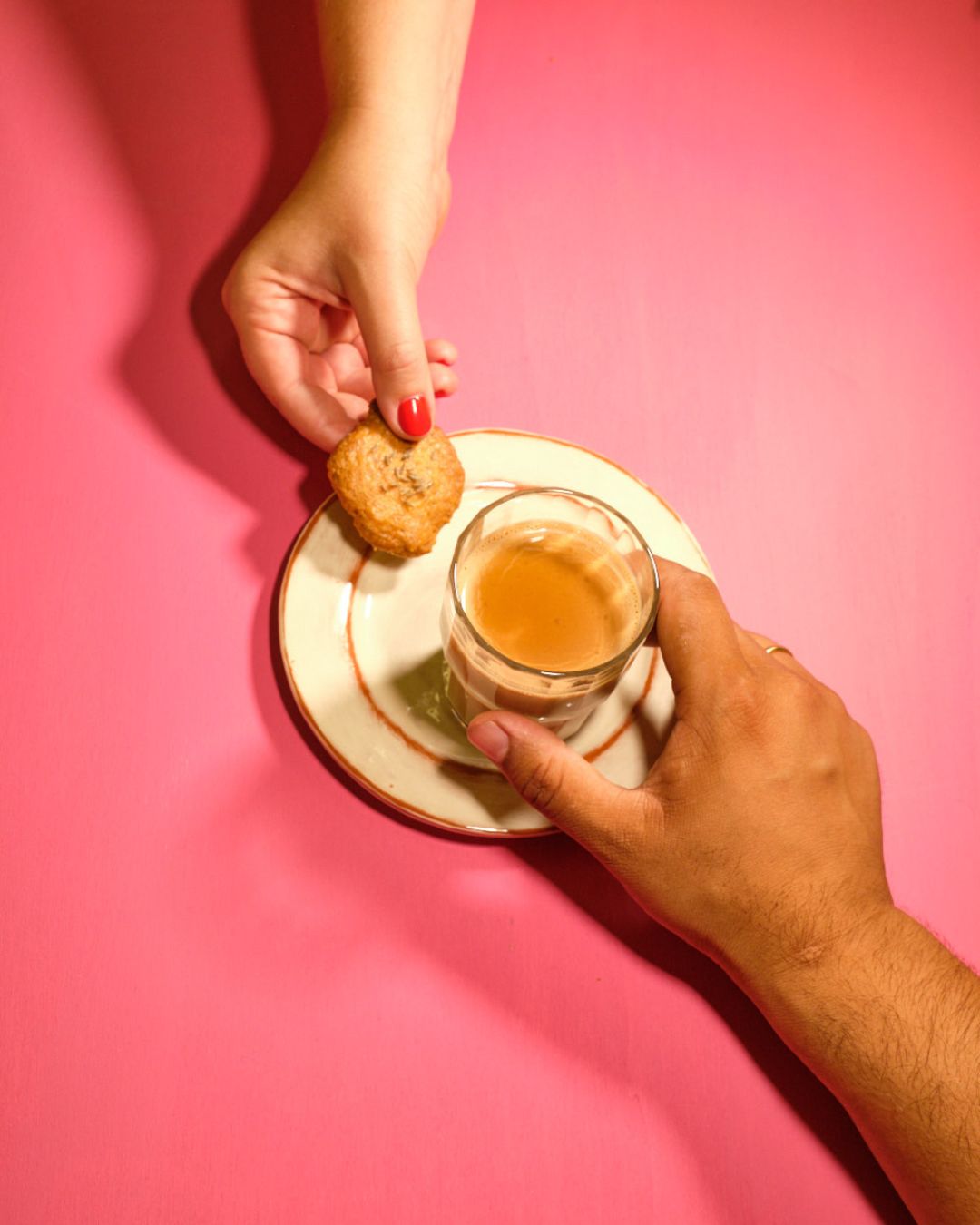 Hand gießt Chai aus einer Metallkanne in einen Becher auf einem blauen Tisch. Daneben ein Teller mit Brownie und ein zweiter Becher Chai. Szene in hellem Sonnenlicht