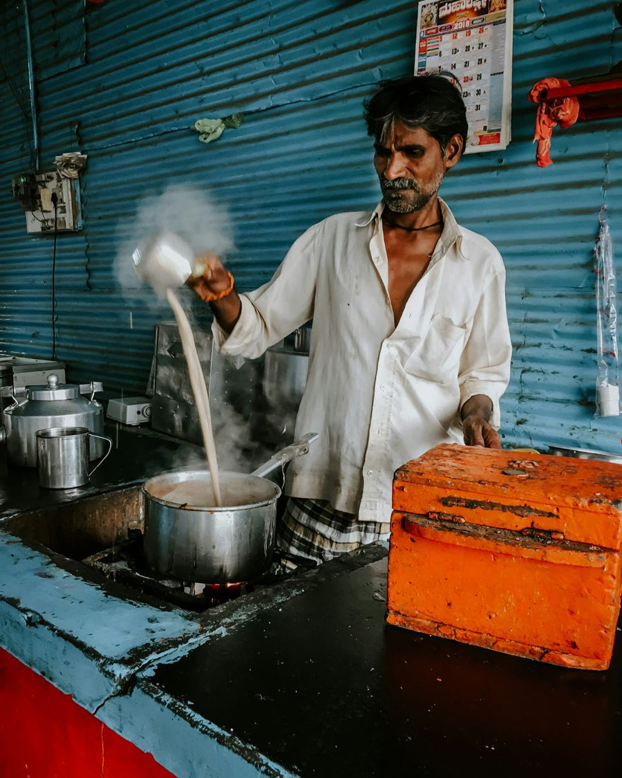 Ein indischer Chai-Verkäufer (Chaiwala) gießt dampfenden Tee aus großer Höhe in einen Kochtopf, um die Aromen zu mischen. Er steht in einer einfachen Teeküche mit blauer Wellblechwand und einer orangefarbenen Kasse