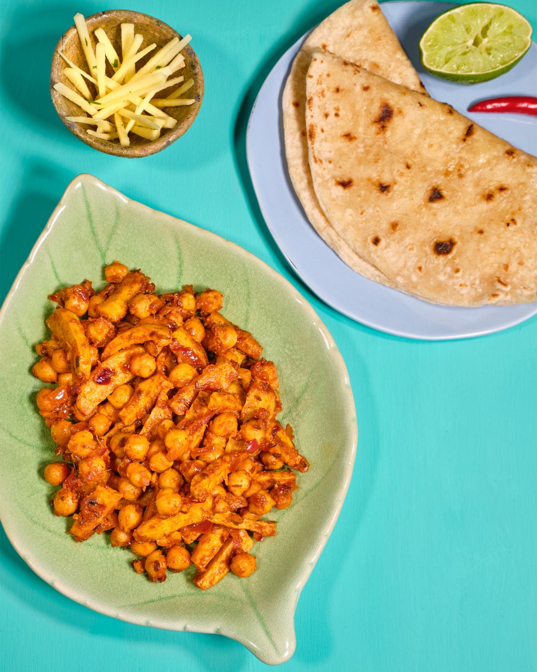 Würziges indisches Kichererbsen-Curry mit Kartoffeln in einer grünen Blattschale, dazu Chapatis, Limette und frischer Chili, Ingwer auf türkisfarbenem Hintergrund
