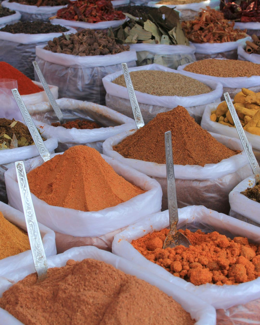 Offene Säcke voller bunter Gewürze auf einem indischen Markt, darunter Chilipulver, Kurkuma, getrocknete Blätter und Samen, jeweils mit Metallschaufeln