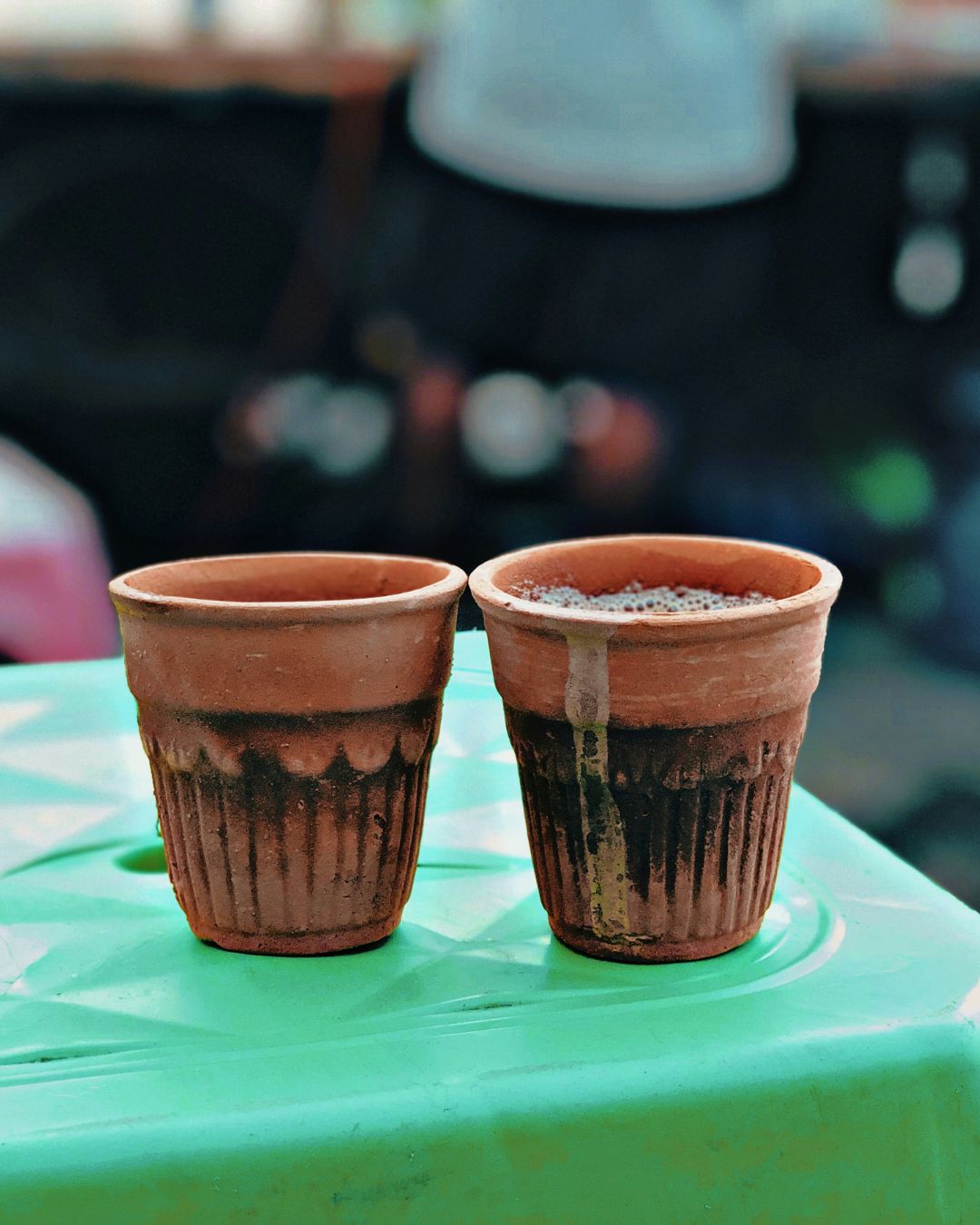 Zwei traditionelle Tonbecher mit frisch serviertem indischen Chai auf einem grünen Tisch. Einer der Becher ist leicht übergelaufen. Im Hintergrund unscharfe Straßenszene