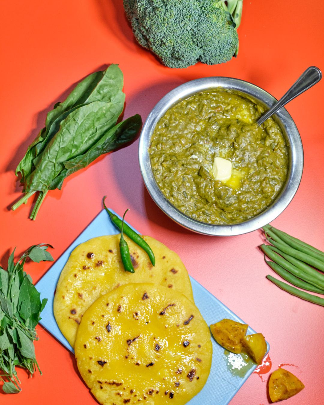Indisches Saag aus Spinat und Brokkoli mit Butterflocken in einer Metallschale, dazu goldgelbe Maisfladen (Makki di Roti) und frische grüne Chilis, angerichtet auf rotem Hintergrund