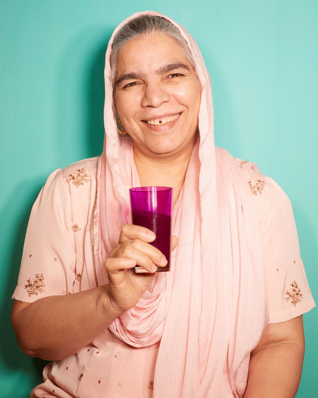 Freundlich lächelnde ältere Frau in rosa traditioneller Kleidung hält ein Glas violettes Getränk vor türkisfarbenem Hintergrund