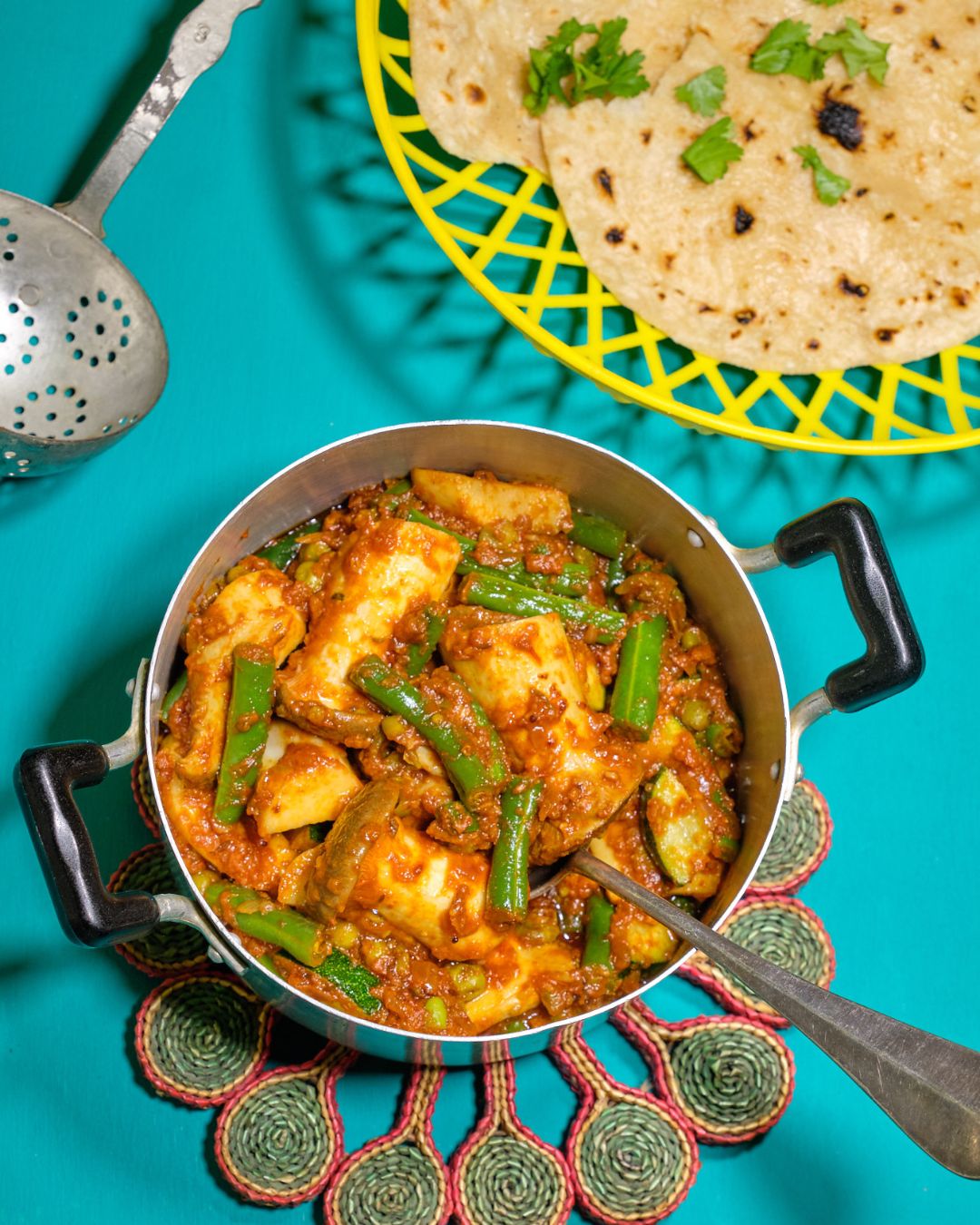 Indisches Pilz-Curry mit grünen Bohnen in einer Metallschale, daneben frische Chapatis auf einem gelben Gitterteller, angerichtet auf einem türkisfarbenen Hintergrund