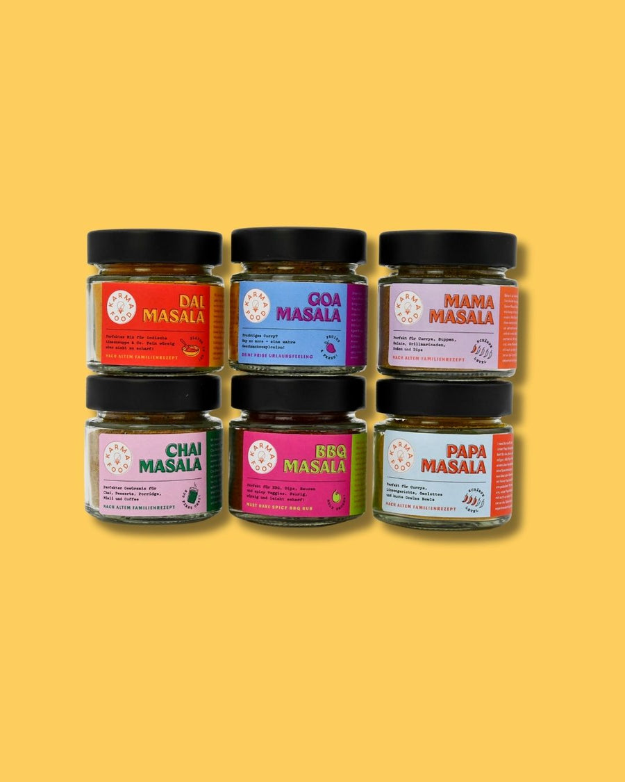 Sechsteiliges Gewürzset von Karma Food mit Dal Masala, Goa Masala, Mama Masala, Chai Masala, BBQ Masala und Papa Masala vor gelbem Hintergrund