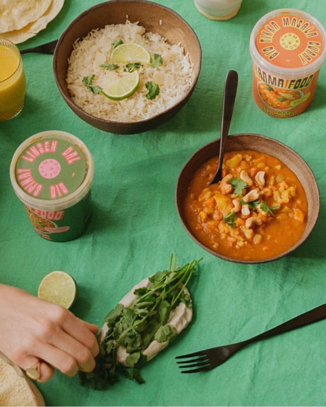 Tischszene mit Bio Spinat Linsen Dal und Bio Tikka Masala Curry von Karma Food, dazu Reis, Koriander und Limetten auf grünem Tuch