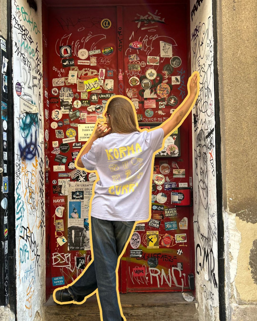 Streetstyle-Look mit Karma Food „Korma Curry“-T-Shirt vor einer Sticker-bedeckten roten Tür in urbanem Setting