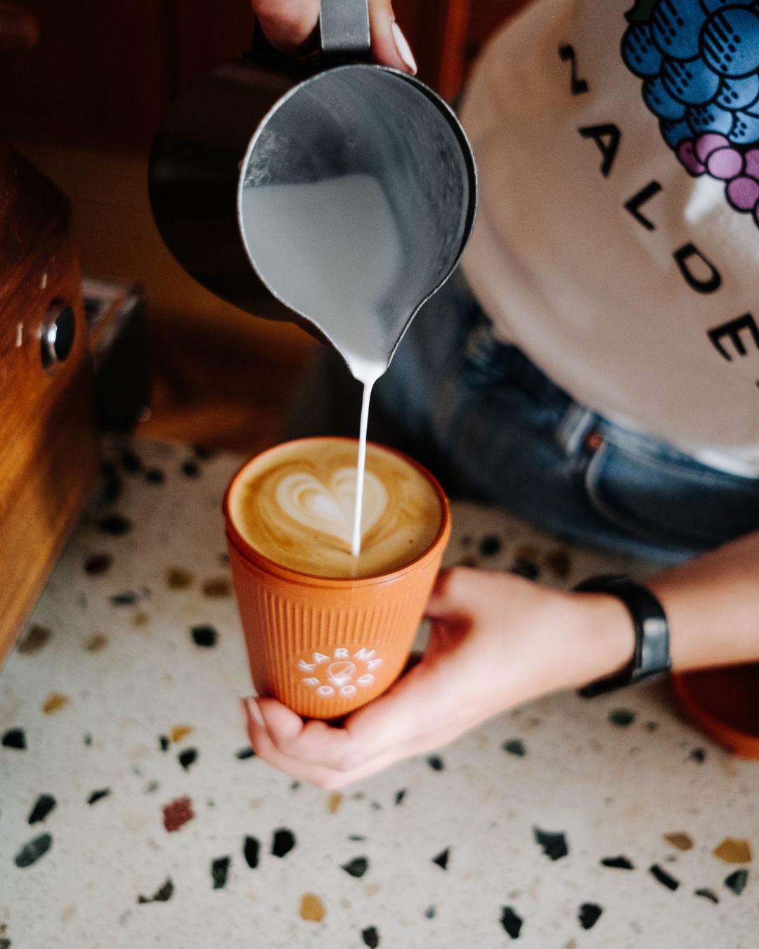 Barista gießt cremigen Milchschaum in einen Latte mit Herzmotiv im orangefarbenen Weducer Cup von Karma Food