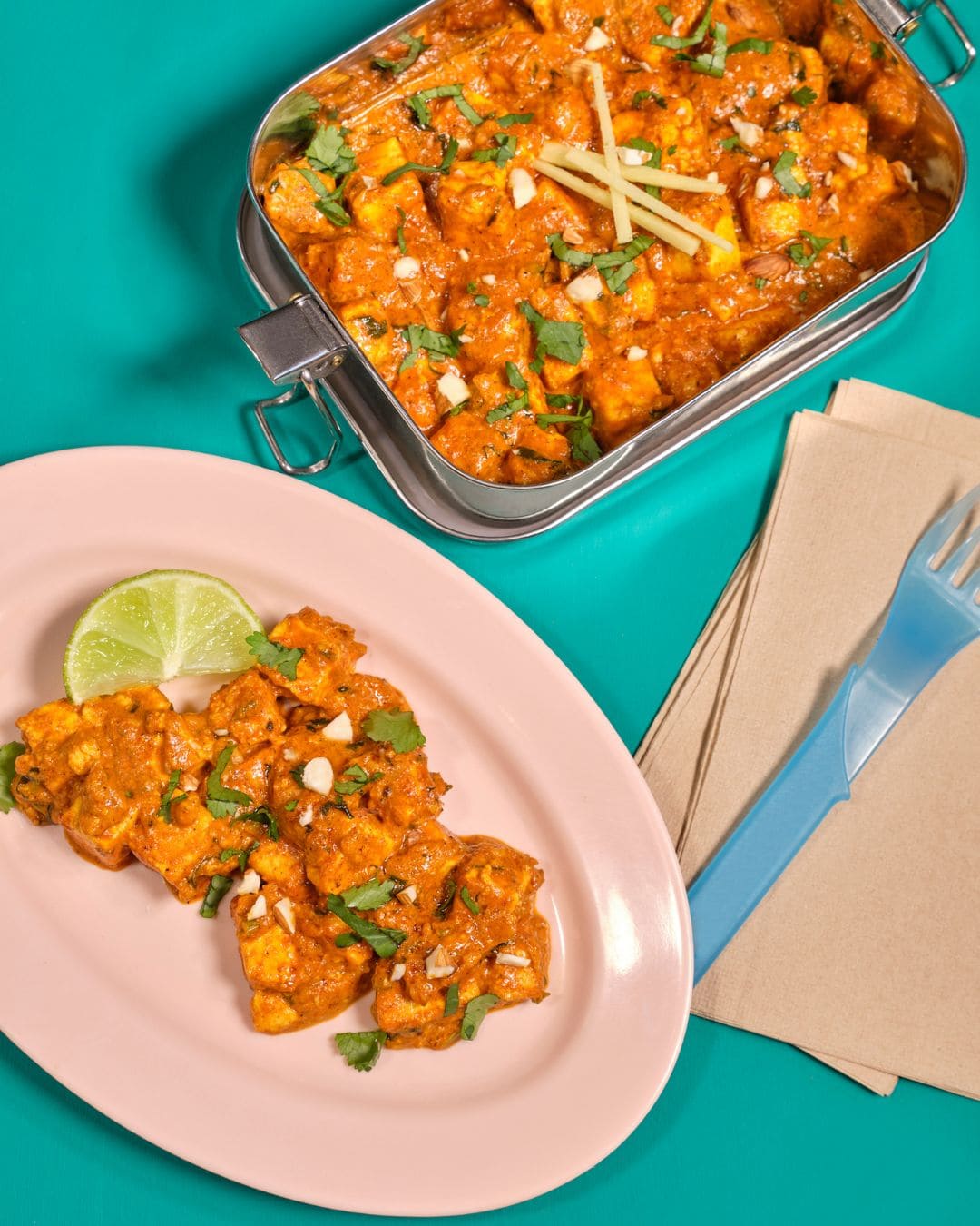 Paneer-Curry in Edelstahlbox und auf einem Teller mit Limettenspalte, angerichtet auf türkisem Hintergrund