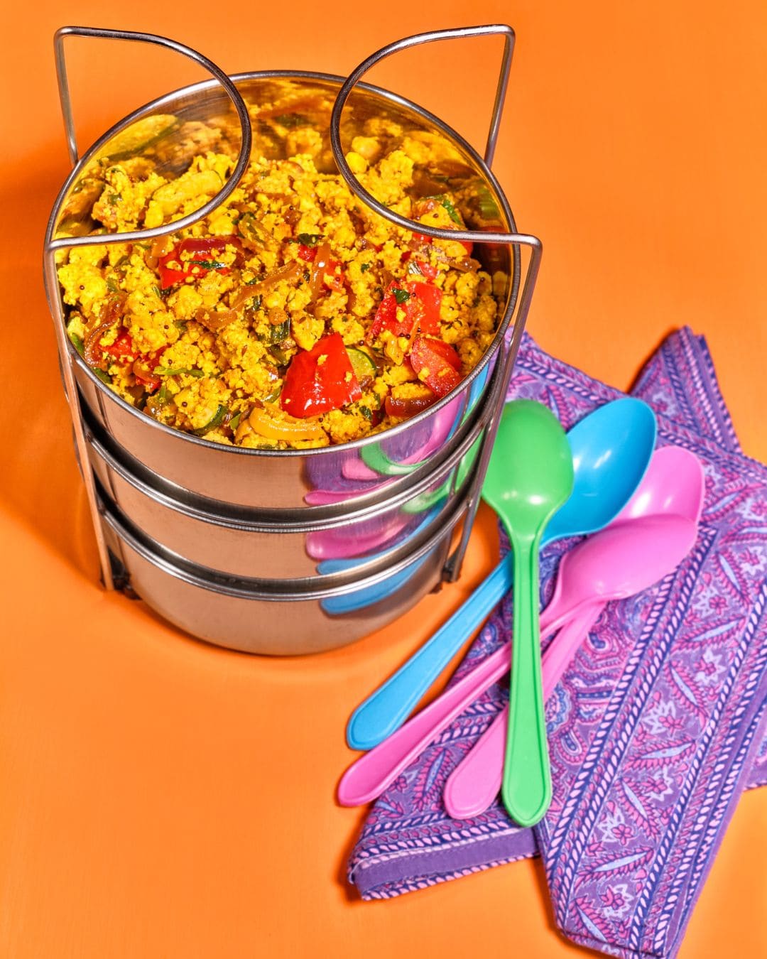 Würziges Scramble-Curry in einer Edelstahlbox, daneben bunte Plastiklöffel auf orangem Hintergrund