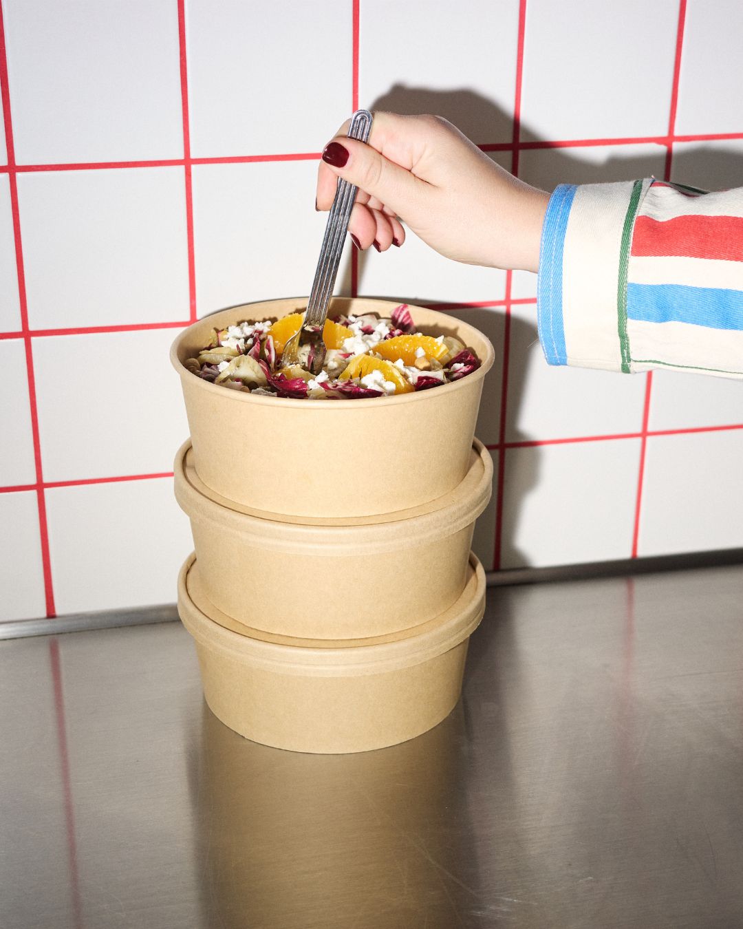 Hand hält Gabel in veganer Bowl mit Orangen und Salat, gestapelte Takeaway-Schalen auf Edelstahltisch vor Kachelwand – veganes Mittagessen in Wien.